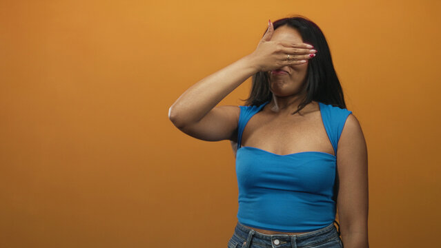 Woman covering eyes with hand, palm over face, wearing blue strap top and jeans, long brunette hair visible against orange studio wall; shyness.