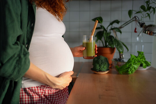 Pregnant woman standing in her kitchen, holding a glass of green smoothie made from fresh herbs and fruits, valuing healthy nutrition and lifestyle choices for maternal and baby wellness