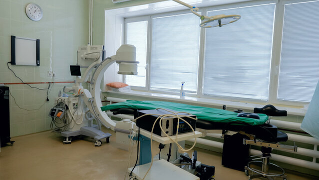 Clean interventional radiology procedure room with adjustable patient table, mobile fluoroscopy imaging arm, ceiling surgical light, monitors, green drape, tiled walls, and daylight through window