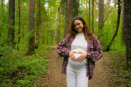 Pregnant woman standing on a forest path, forming a heart shape with her hands on her baby bump, smiling and feeling happy, symbolizing new beginnings and awaited arrival