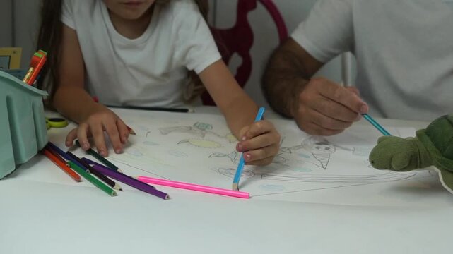 Detailed macro shot of a young child's hands holding a blue pencil and carefully coloring a cute narwhal drawing on a white sheet of paper.
