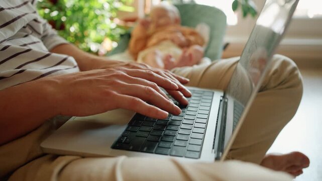 Young freelancer dad is typing on his laptop computer while caring for his infant child in a bouncer at home, illustrating the challenges of combining remote work with parenthood