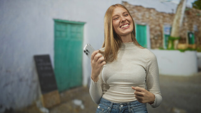 Woman holding smartphone, smiling and pumping fist while later speaking on phone on a street with green doors and stone walls of a village; celebration joy.