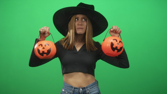 Woman witch wearing wide brim hat holds two jack o lantern pumpkin buckets by raised hands showing bare midriff against green screen studio backdrop; skeptical halloween fun.