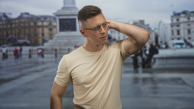 Man with glasses touching his head with right hand in a wet city street by a monument and buildings; thoughtful introspection.