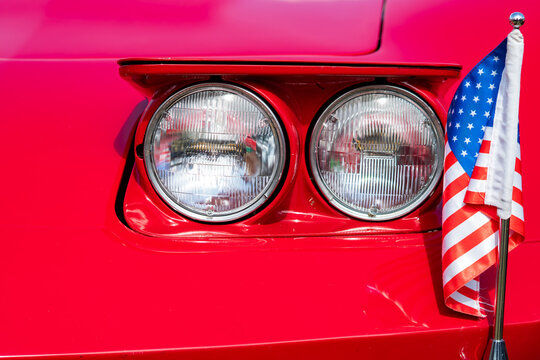 Close-up of the front of a vintage red sports car with round, dual headlights and a small United States flag attached to the bodywork, intense lighting, saturated color, classic automotive.