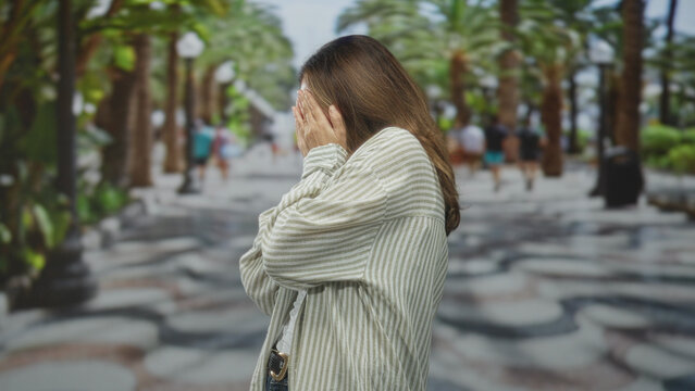Woman with hands covering face on a palm lined street, wearing striped shirt and jeans, side profile visible; anxiety vulnerability.
