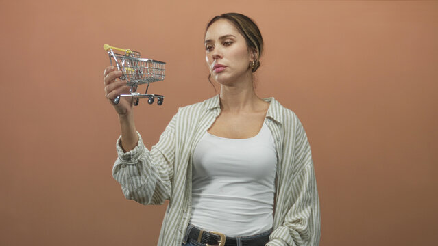 Woman holding miniature shopping cart by hand in studio, inspecting it with pensive gaze; contemplation thrift budgeting.