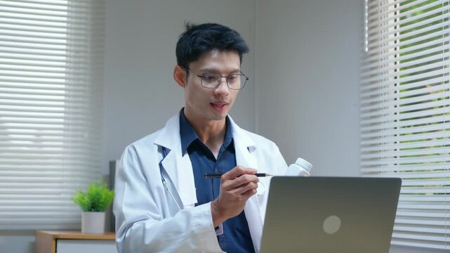 Asian male doctor providing online consultation to patient using laptop and showing medicine bottle in his modern office
