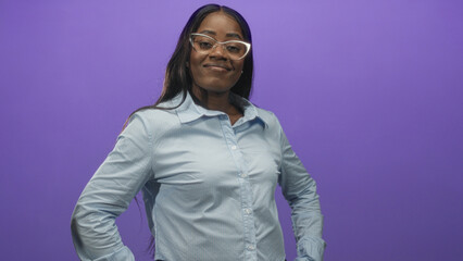 Obraz premium Woman wearing glasses and a light blue buttoned shirt, hands on hips in studio with purple backdrop; confidence poise.