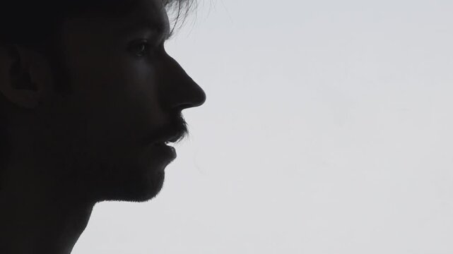 Silhouette profile of young man exhaling thin vapor plume against pale backlit background, tight sideview focusing on head and upper neck with negative space filled by dissipating cloud