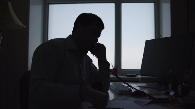Silhouetted office worker speaking on telephone and taking notes at desk with strong backlit window