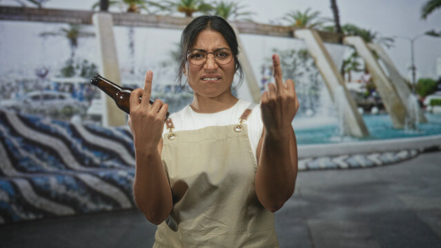 Woman holding beer bottle and flipping middle finger on a street plaza with fountain and mosaic wall; defiance boldness.