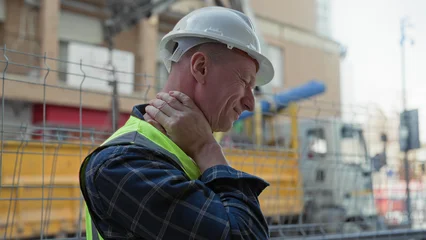 Man builder in white hardhat and neon hi vis vest holding neck with hand, grimacing beside construction fence and truck at building site  pain safety. © Krakenimages.com