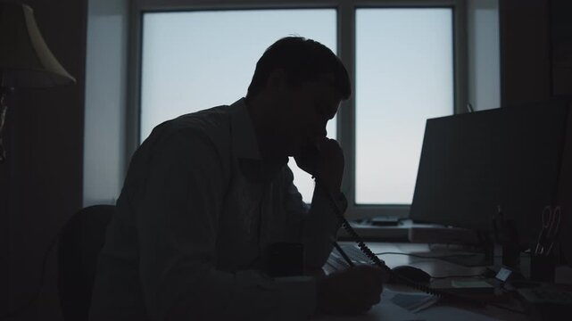 Silhouetted office worker holding corded phone beside large desk in backlit workspace with window glow, focused professional mood, serious posture, modern corporate interior