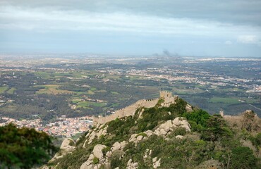 Ancient castle walls cling to a rugged hilltop in Sintra, Portugal, overlooking vibrant green fields and quaint towns. Nature and history intertwine in this striking panoramic view © Damian