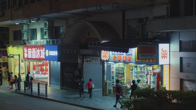 A lively Hong Kong street scene at night. The view is an elevated street-level perspective looking towards a row of brightly illuminated storefronts