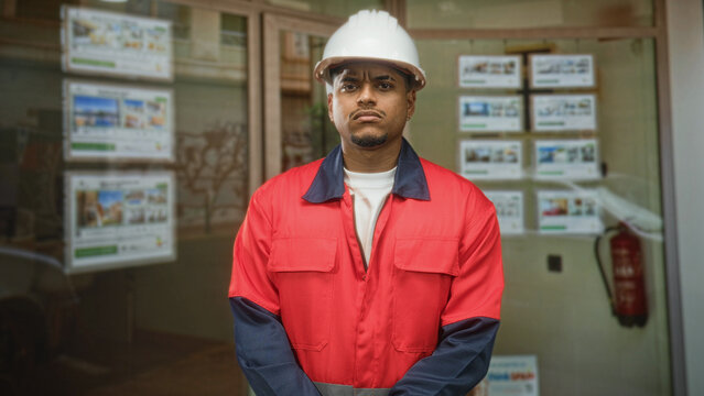Man with beard in white hard hat and red coverall looks left at storefront in building; quiet determination.