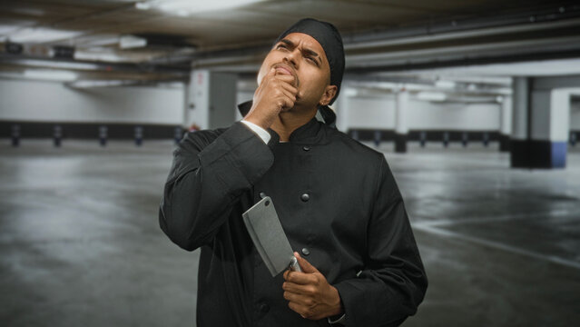 Hispanic man chef in black uniform holding cleaver, hand on chin, in parking garage; thoughtful pondering.