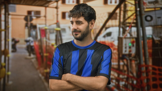 Young hispanic man with beard in blue football shirt and team jersey, arms crossed and pouting while standing by construction scaffolding on a street; disappointment.