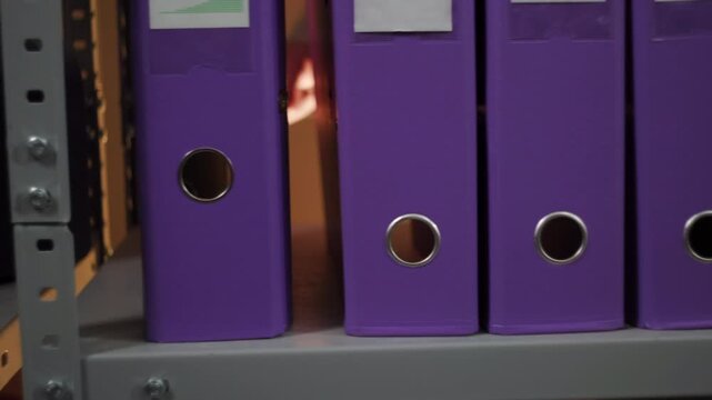 Upright purple binders on office shelf with one binder slightly pulled forward exposing narrow gap, circular finger holes, cardboard edges, neat storage, muted light, orderly side view