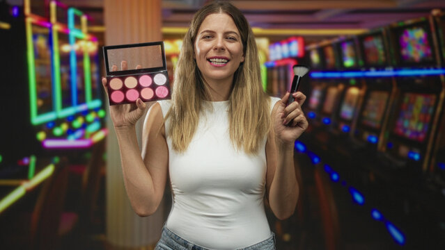 Woman holds makeup palette and brush in a casino gaming building, smiling and showing the cosmetics; beauty tutorial confidence.