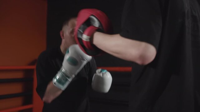 Male boxer training with partner mitt work in dim indoor gym, guarded stance and landing punch with visible sweat and motion blur