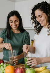 Obraz premium Two smiling women preparing healthy green smoothies and flax seeds in a modern kitchen, wellness and nutrition concept, Fresh fruits and vegetables are visible on the wooden countertop