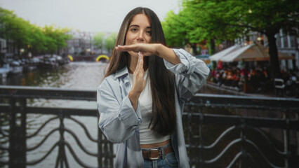 Fototapeta premium Woman makes time-out t gesture with hands on street canal bridge by riverside railing and outdoor cafe; confidence.