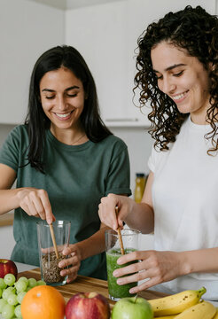Two smiling women preparing healthy green smoothies and flax seeds in a modern kitchen, wellness and nutrition concept, Fresh fruits and vegetables are visible on the wooden countertop