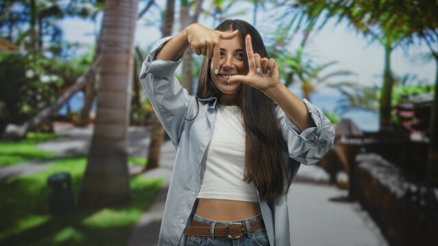 Woman framing face with hands on a beachside promenade street wearing crop top and jeans; playful confidence.
