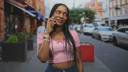 Fototapeta premium Black woman holds phone to ear, smiling with braided hair and visible midriff in a pink crop top on an urban street; happy confidence.