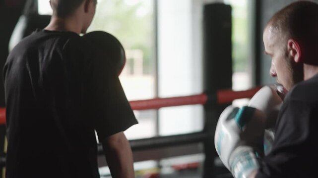 Boxing pad work in gym ring with trainer holding focus mitt while boxer drives punches forward under bright window backlight and gritty brick wall atmosphere, with boxing, trainer, boxer, mitt detail
