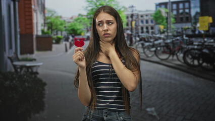 Fototapeta premium Teenage girl holds small red heart and clutches her throat while standing on a city street with parked bicycles and buildings; heartbreak.