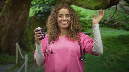 Fototapeta premium Woman with curly hair in pink scrubs holding a blue pill bottle, stethoscope around neck, smiling and waving hand in forest path; care hope.