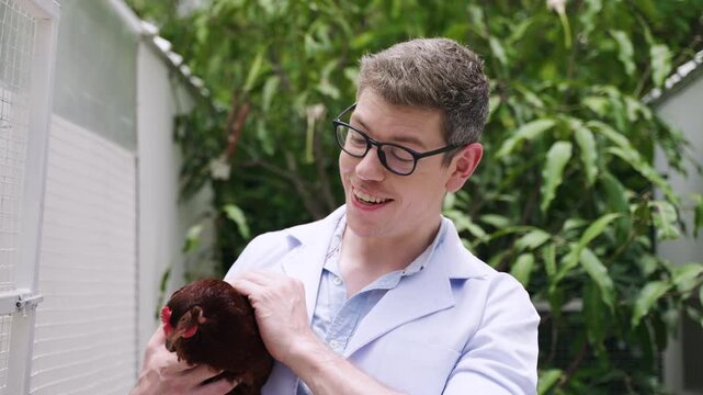 Poultry veterinarian handsome Caucasian man wearing eyeglasses standing holding a hen for health checking with smiling and happiness in the henhouse at an organic farm. 