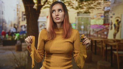 Fototapeta premium Woman in yellow sweater holding a pair of mustard socks by the cuffs and showing them with a puzzled expression on a street cafe; uncertainty.