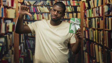 Man holding green gift box with white ribbon while giving thumbs down and frowning amid bookshelves in a building  disapproval rejection. © Krakenimages.com
