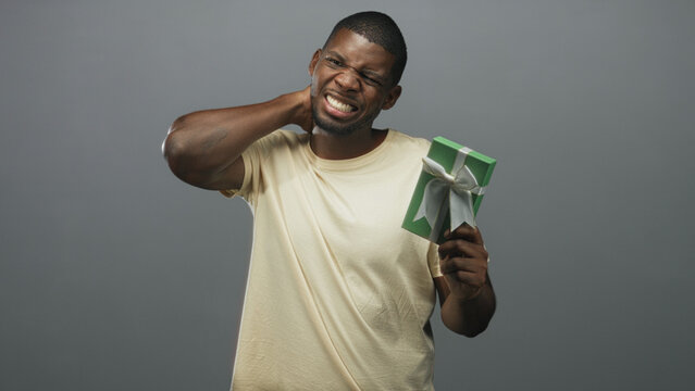 Young black man holding a green gift box with ribbon, rubbing his neck and grimacing in a gray studio; surprise pain.