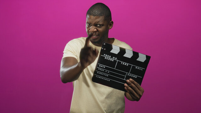 Young black man holding clapperboard and pointing finger at camera in studio; confidence assertiveness.