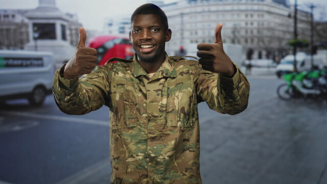Man soldier in camouflage giving thumbs up, hands raised and smiling on a busy urban street with vehicles and a monument visible; pride duty.