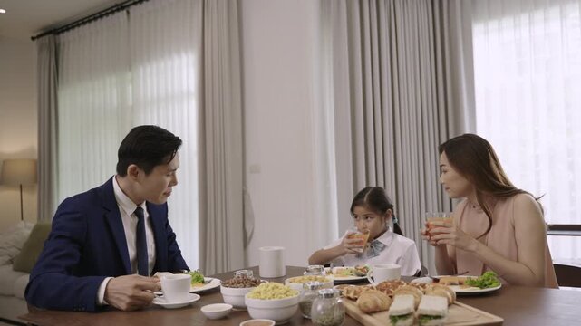 Happy Asian family father, mother, and daughter sitting smiling while eating breakfast and drinking orange juice for health together in morning in kitchen at home. Dad in suit, girl in school uniform.