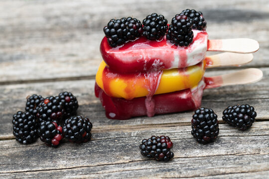 Three popsicles are stacked on top of each other on a rustic wooden board. The popsicles are starting to melt. There are blackberries on top of and next to the popsicles.