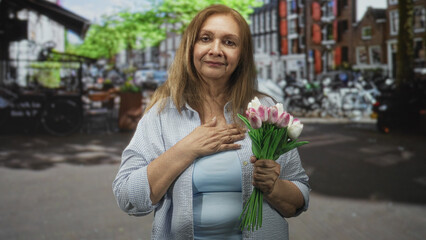 Fototapeta premium Woman holding pink and white tulips, hand over chest, standing on a city street with bicycles and brick canal houses; gratitude reflection.