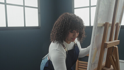 Woman painting on canvas in a studio, side profile wearing apron, arm reaching with paintbrush at easel near window light  quiet concentration. © Krakenimages.com