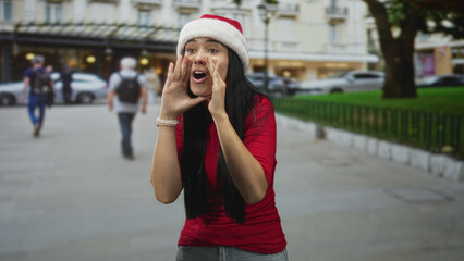 Fototapeta premium Young woman in a santa hat cups her hands to mouth, points and signs with both hands while speaking on a busy city street in front of shops and parked cars; festive.