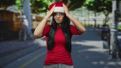 Obraz premium Young hispanic woman holding santa hat with both hands on head on a street lined with trees and market stalls, wearing red top and long hair; holiday anxiety.