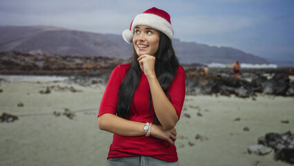 Obraz premium Young woman wearing santa hat smiles with hand on chin and crossed arm in a studio beach set; holiday joy.