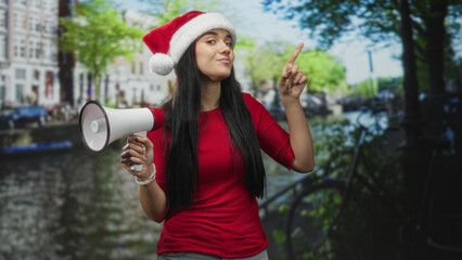 Obraz premium Young woman wearing santa hat holds a megaphone and points finger on a city street by canal buildings, smiling and addressing unseen crowd; holiday cheer.