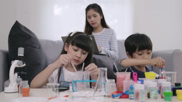 Happy Asian boy and girl students are sitting study learning science and conducting chemistry experiments together with a young teacher sitting and taking care in the back of a nursery classroom lab. 